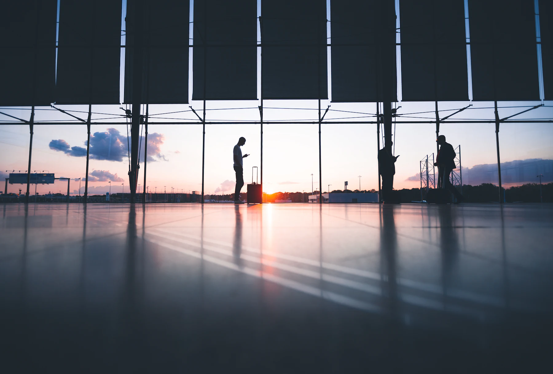 Travellers silhouetted against a low sun behind the tall windows of an airport terminal, a suitcase standing by the glass