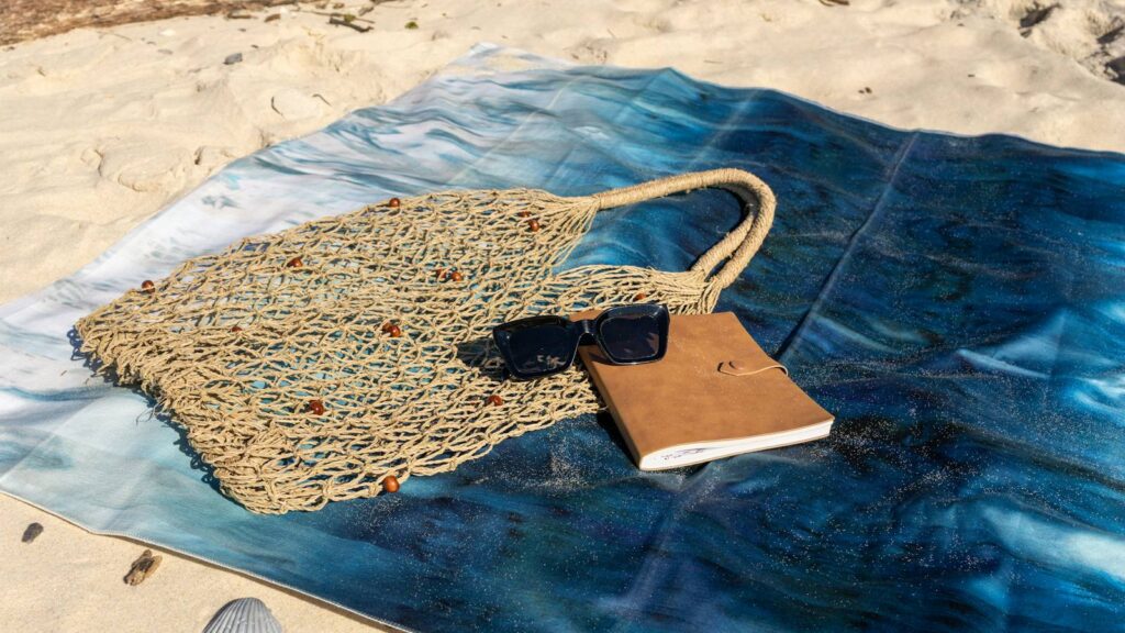 A woven string bag, sunglasses and a notebook lying on a blue patterned beach towel on the sand