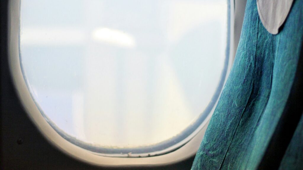 A softly lit aircraft window beside a teal patterned passenger seat with a white headrest cover