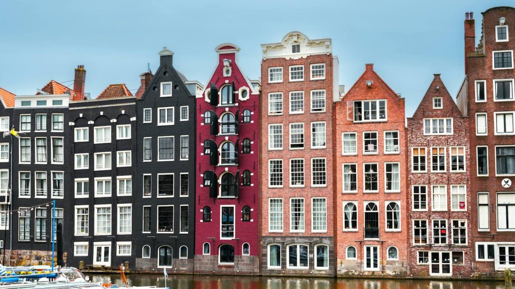A row of tall, narrow canal houses in black, plum and red brick reflected in the water below