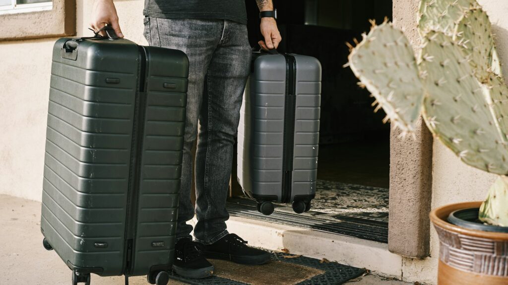 A traveller in jeans standing at a front door holding a large and a cabin-size hard-shell suitcase