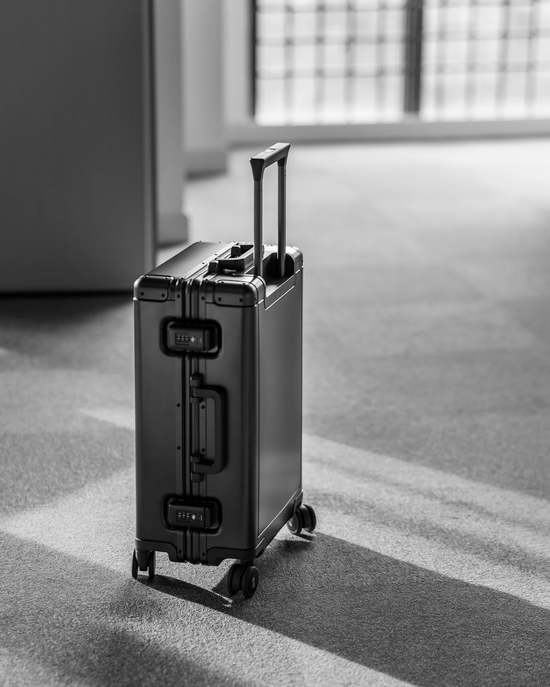 A black-and-white photo of an aluminium carry-on suitcase with its handle raised, standing on carpet in window light