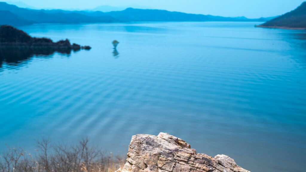 A calm blue lake surrounded by hazy hills, seen from a rocky outcrop