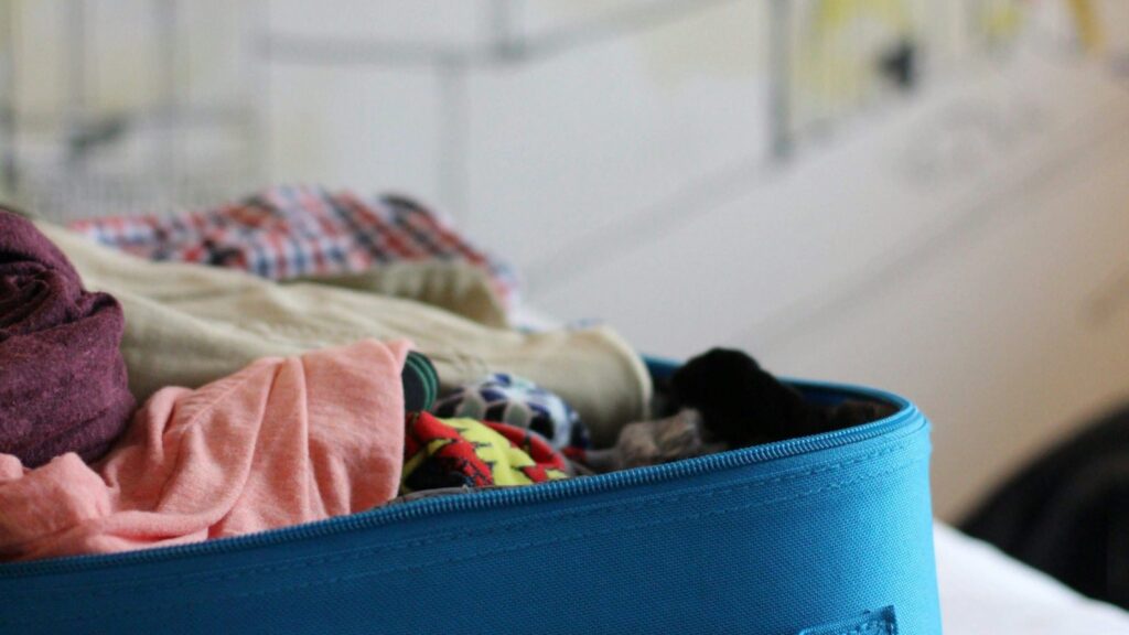 An overfilled blue soft suitcase with folded and crumpled clothes spilling over the zip