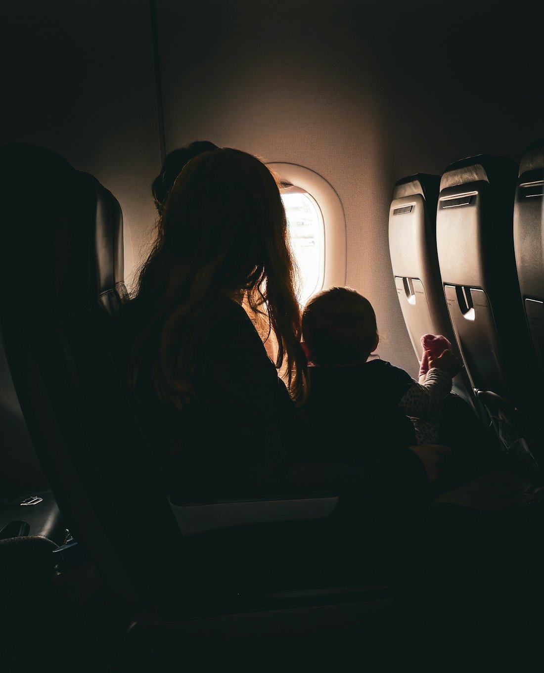 A parent and a small child in silhouette, looking out of an aircraft window from their seats