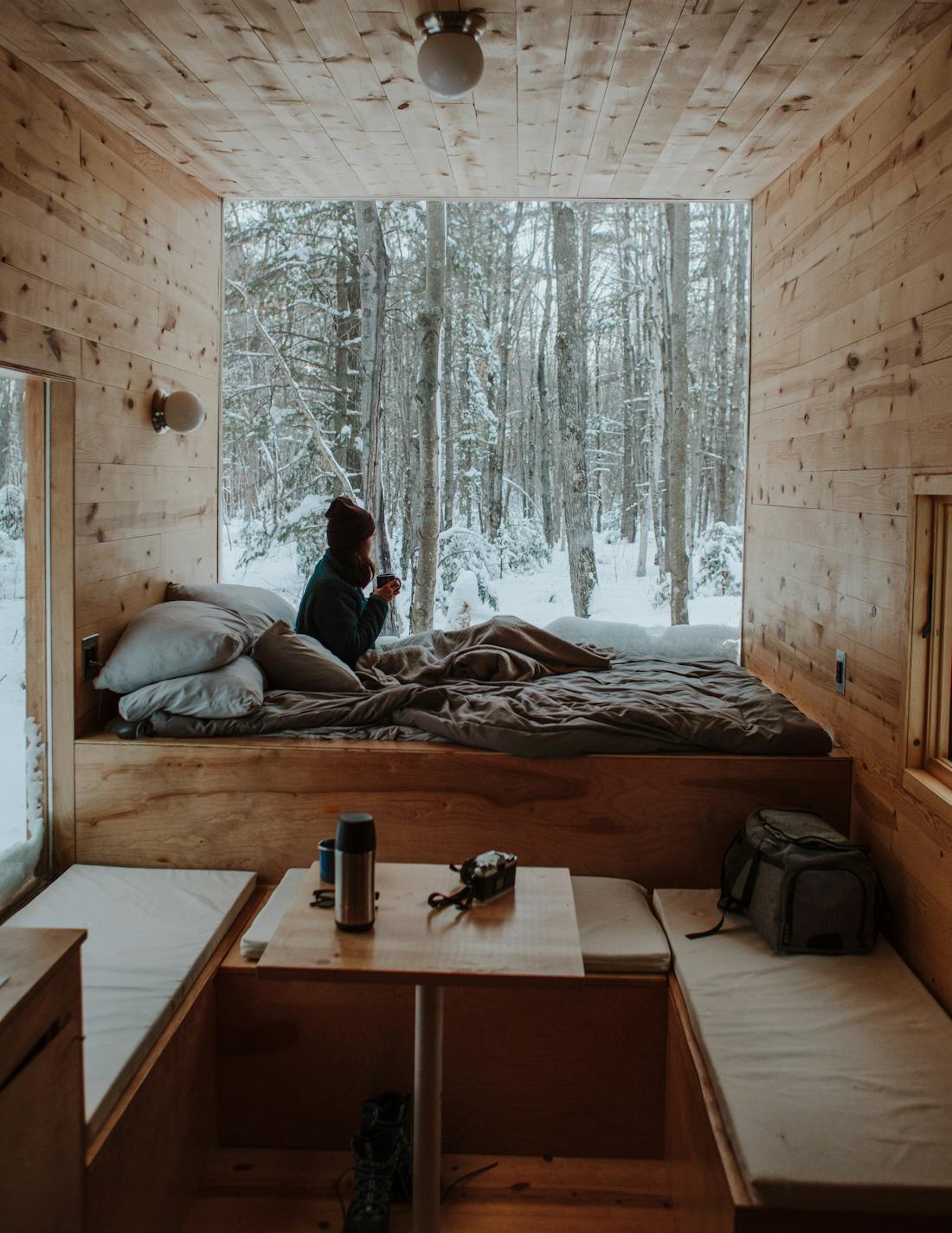A traveller in a woollen hat sitting on a bed in a wooden cabin, looking out at a snowy forest