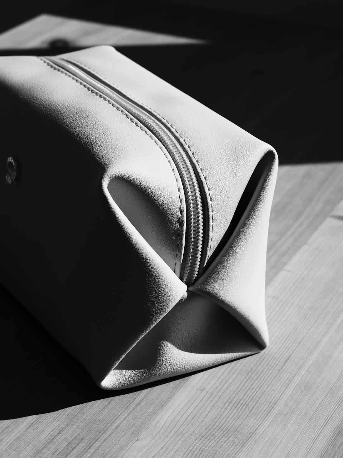 A black-and-white close-up of a zipped leather toiletry pouch on a wooden surface in strong window light